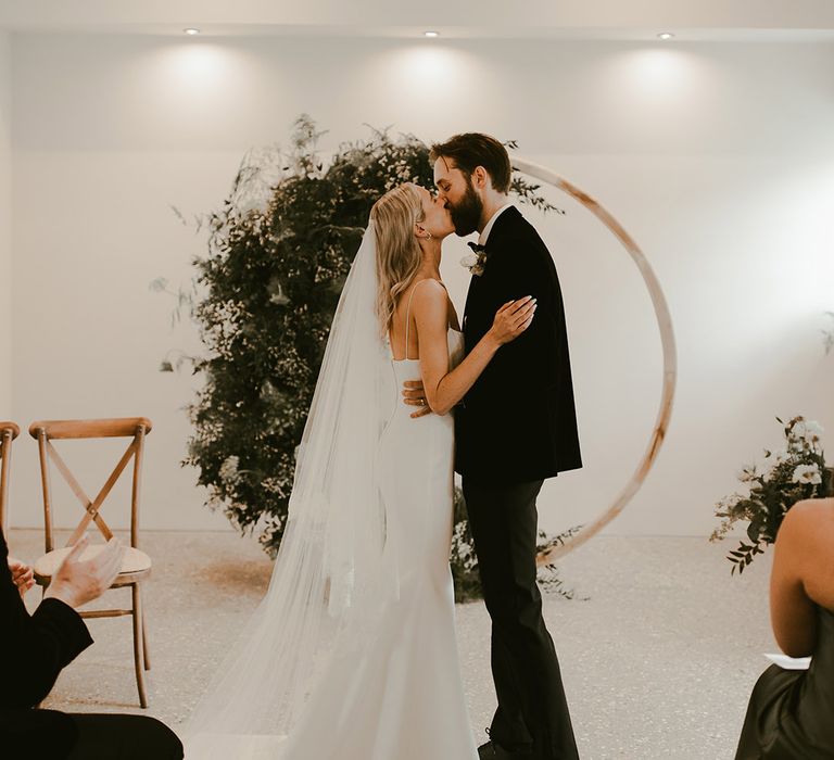 Groom in black tie and bride in fitted wedding dress with thin straps she their first kiss as a married couple in front of the moongate decorating the altar