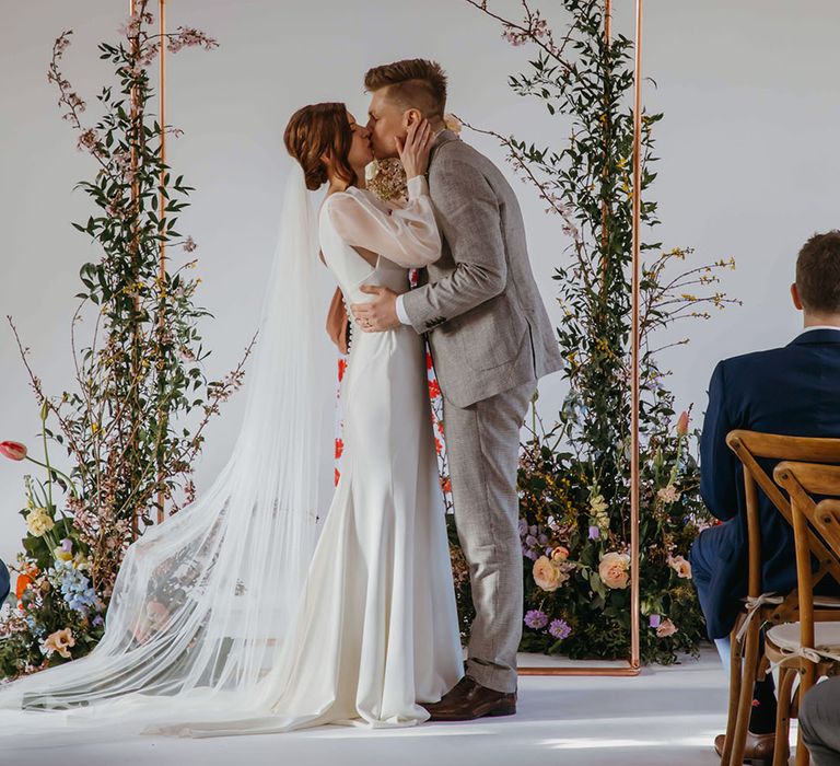 Bride and from share their first kiss as a married couple with a copper frame and wildflower flower column for stylish wedding 