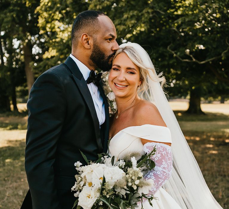 Bride in drop veil and carrying white floral bouquet leans in to her groom in black-tie 