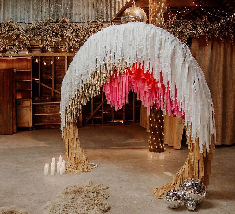 Boho barn wedding venue with fairy lights, pillar candles, disco ball wedding decorations and a large semi-circle arch decor with white, gold and hot pink streamers - perfect streamer decoration ideas 