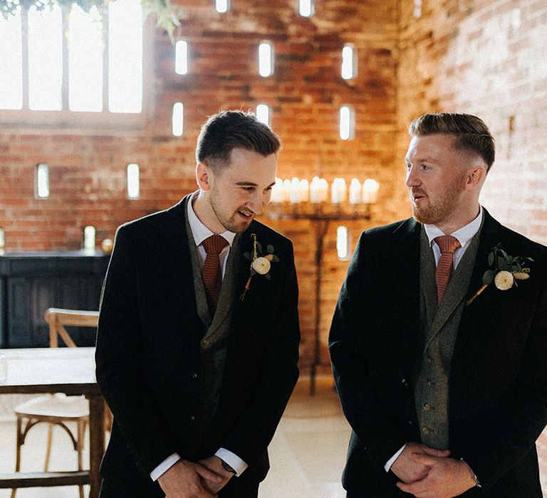 The groom and his best man stand talking at the altar wearing matching suits with orange ties and white flower buttonholes