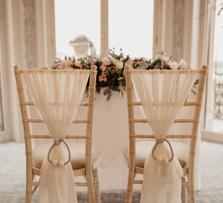 Light wooden wedding chairs decorated with white sash tied with rope and pearls 