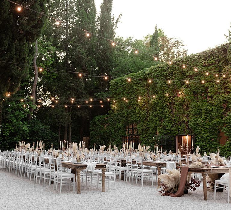 Wooden banquet tables for outdoor destination wedding reception with neutral styling, pampas grass arrangements and macrame decor