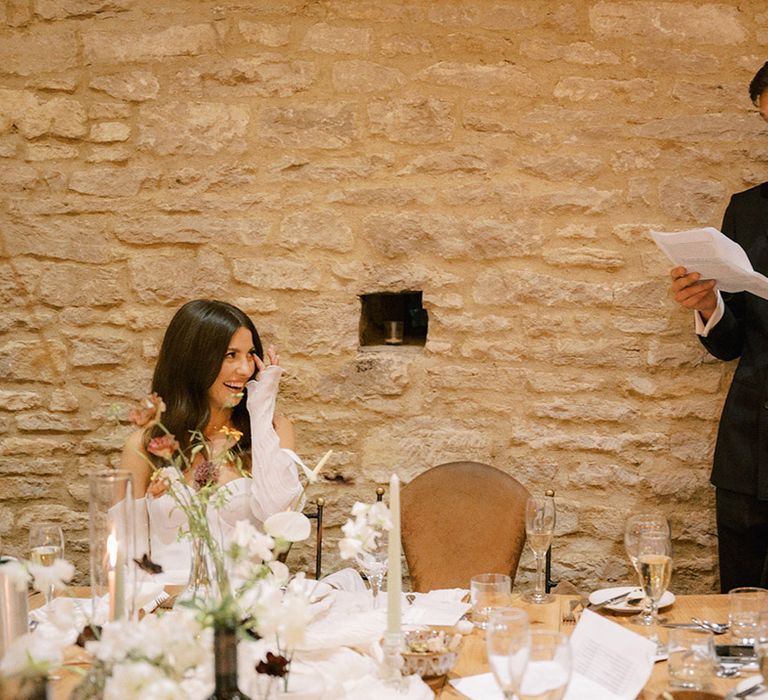 Groom in dark double breasted navy suit standing up reading out his groom speech with the bride sitting down enjoying the speech at the reception 