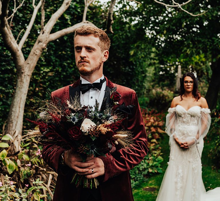 Bride in off the shoulder lace wedding dress with puff sleeves waits behind the groom who wears a burgundy velvet tuxedo holding the bouquet