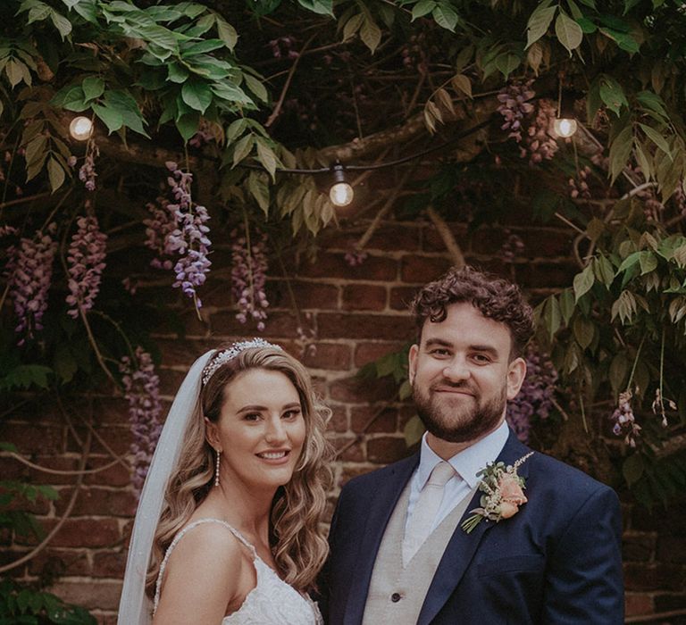 The bride wears a lace fitted gown holding a peach and white bouquet standing with the groom in a navy suit jacket and beige waistcoat with a pale tie