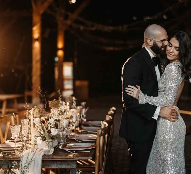 Bride in open back silver sparkly wedding dress and groom in classic black tuxedo standing beside contemporary modern luxe wedding tablescape at Elmley Nature Reserve 