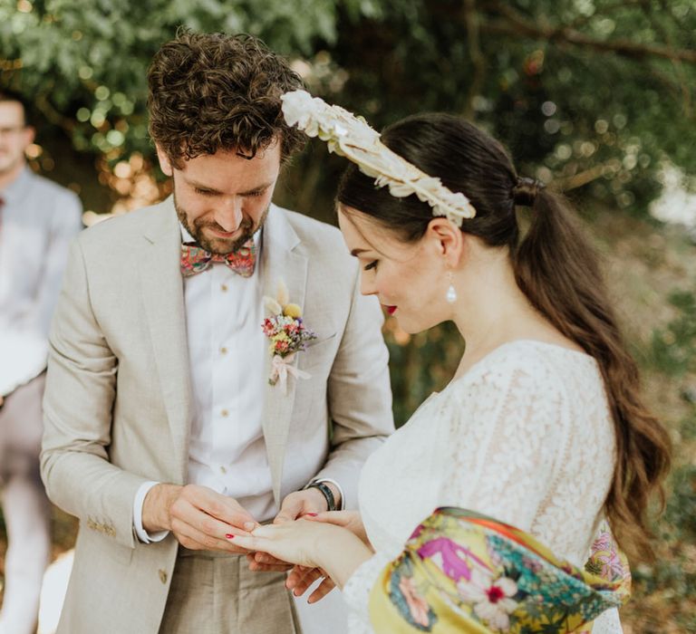 Bride in white lace wedding dress and boho bridal vrown exchanges rings with groom in greige suit and colourful bow tie