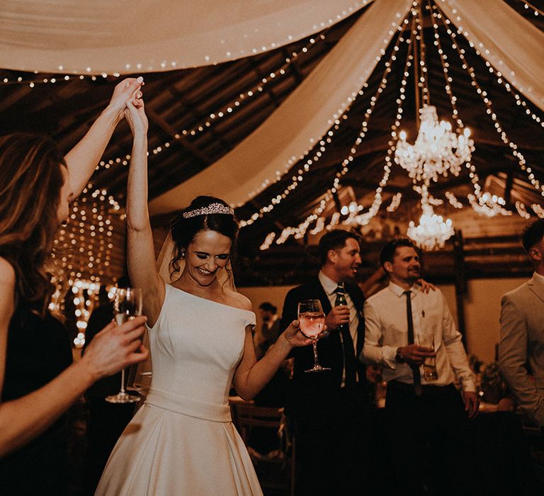 Bride in off the shoulder wedding dress with tiara dances with wedding guest at the reception lit up with cosy fairy lights 