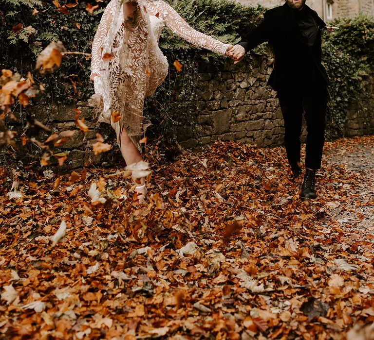 The bride in a Catherine Deane lace wedding dress and groom in all black wedding outfit play out in the autumn leaves