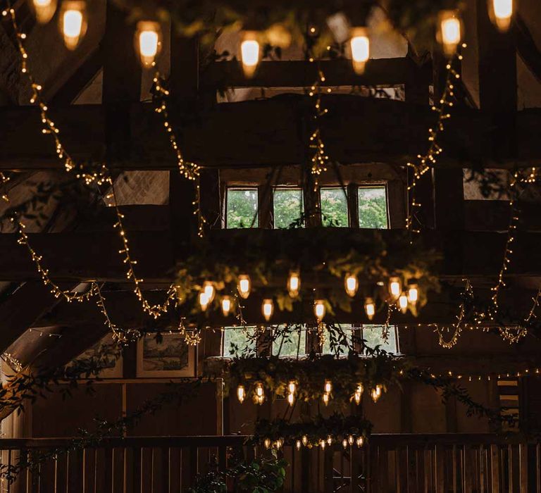 Bride and groom standing in Lains Barn reception room with fairy lights, suspended foliage arrangements, festoon chandeliers, large neon wedding sign, foliage wall and candles 