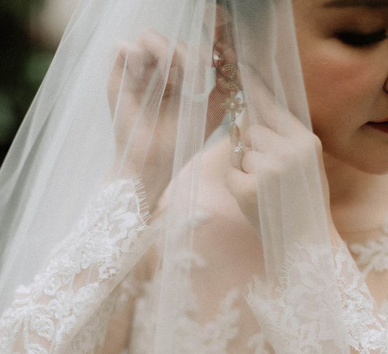 Bride in long sleeve lace wedding dress with delicate white veil wearing Chanel pearl and diamond earrings 