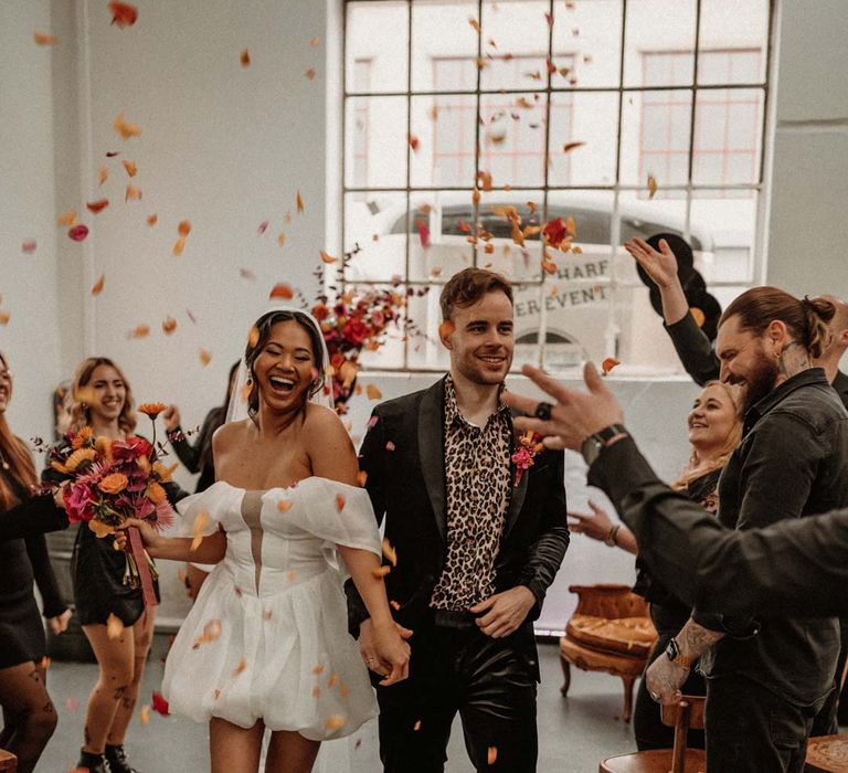 Groom in wet look black grooms suit with leopard print shirt and pink and orange boutonniere standing by vinyl wedding arch walking hand in hand with bride doing colourful wedding confetti exit with bride in off shoulder tulle short wedding dress with church length veil and white platform Dr Martens wedding shoes