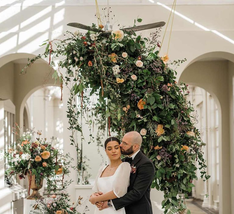 Groom in classic black tuxedo with bowtie and wildflower boutonniere dancing with bride in short sheer puff sleeve wedding dress with square neck and wrist loop with sheer pearl embellished bridal veil surrounded by suspended wildflower and foliage arrangements in large gold vases at Buxted Park