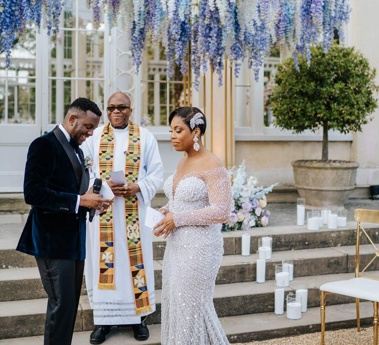 Bride in illusion v-neck sheer off shoulder long sleeve pearl embellished wedding gown standing with groom in groom in velvet midnight blue double breasted groom’s blazer, velvet bowtie and peach garden rose boutonniere underneath suspended pastel flower arrangements at Syon Park vow renewal 