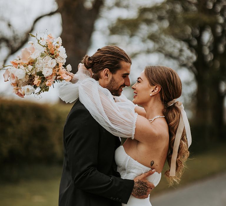 Bride in corset style wedding dress with detachable sleeves and pearl jewellery kissing groom in black tie with neutral wedding bouquet 