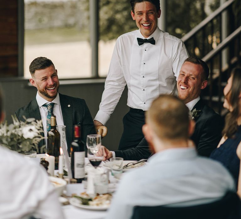 Groom wearing white shirt and black bow tie stands socialising with the wedding guests 