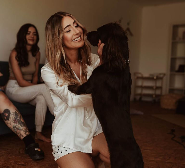 Bride wearing white satin pyjamas petting her dog as she gets ready for the wedding morning 