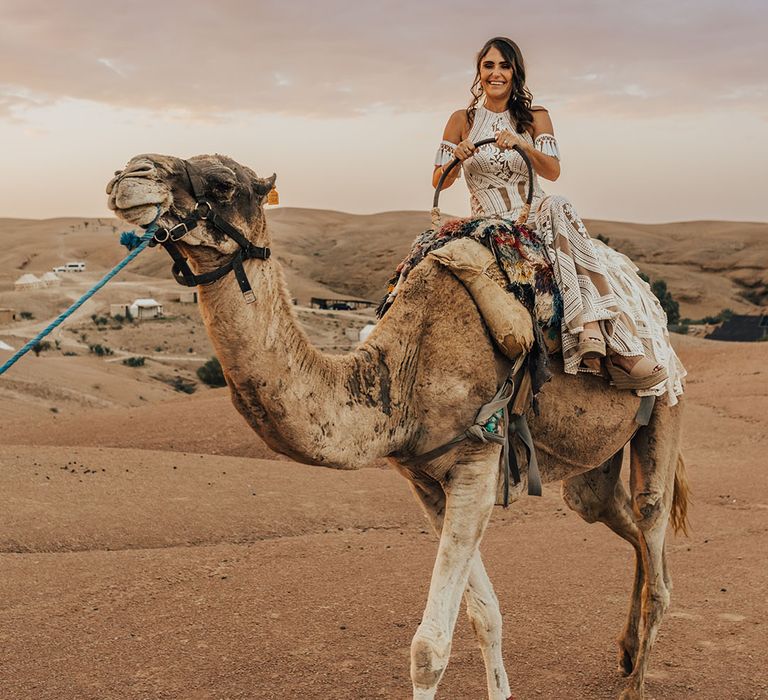 The bride in a boho lace tassel wedding dress rides upon a camel at destination wedding 