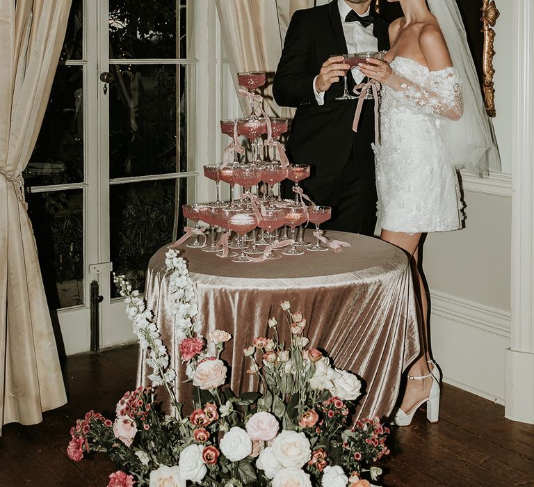 Bride in short wedding reception dress with groom in black tie next to pink champagne tower decoration with velvet bows 