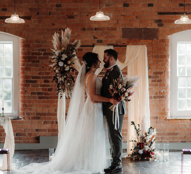 The bride and groom stand in front of the boho altar decoration made from white drapery and dried flower arrangements on wooden frame 