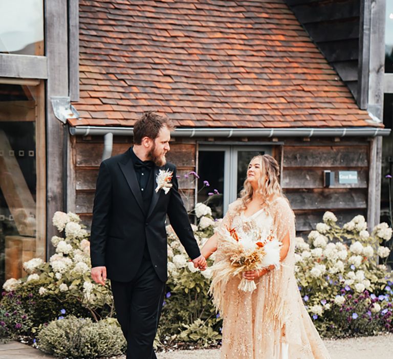 Groom in all black wedding suit walking with the bride in a gold wedding dress for their romantic couple portraits 