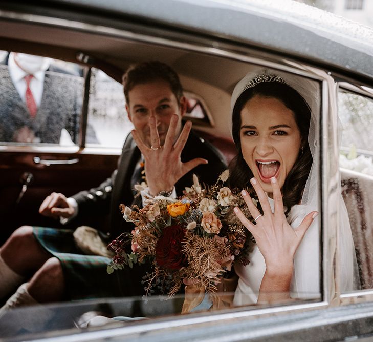 Bride holds up her hand to show her wedding ring from inside a wedding car. She wears a veil and has brunette hair.