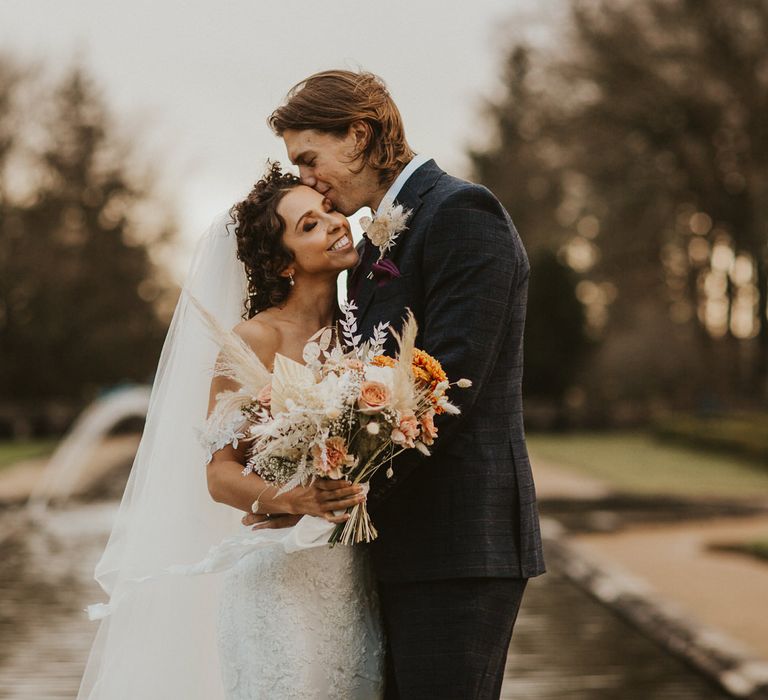 Groom in a navy suit kissing his brides head in a lace wedding dress and cathedral length veil holding a neutral, fresh and dried flower wedding bouquet 