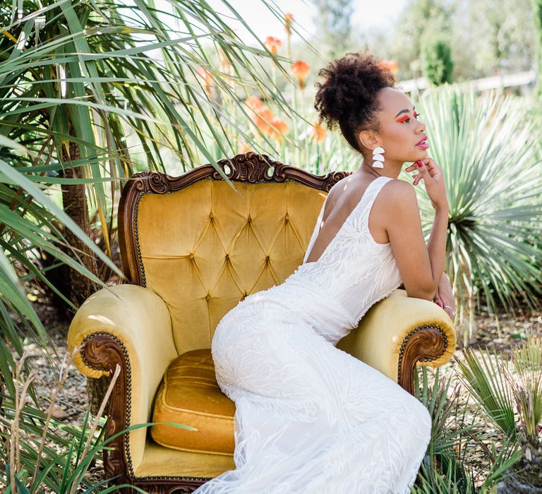 A bride sits on a yellow chair. She has a low back white dress on with statement white earrings. She has bold orange eyeshadow and wears her curly hair high on her head