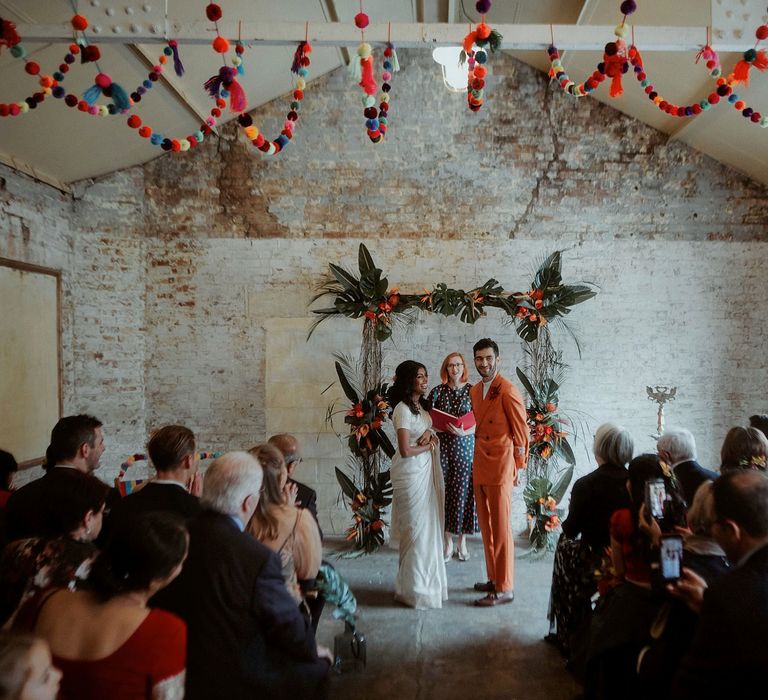 Bride in a white sari and groom in an orange suit at their industrial wedding with colourful pompom ceiling decor and tropical plant altar. 