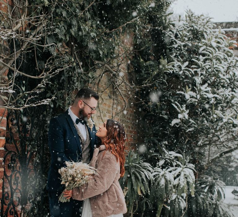 Bride in fur coat holding dried flower bouquet looks up at groom in navy suit in the snow 