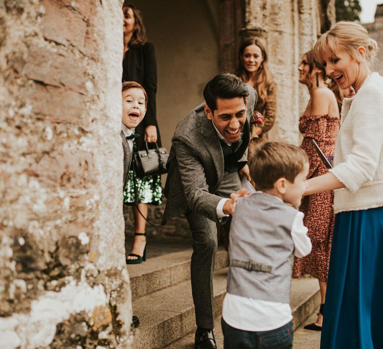 Groom in a grey wool suit laughing with a little wedding guest outside the church 