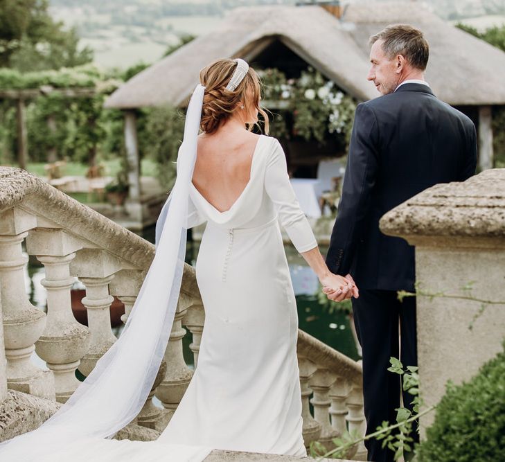 Bride in cowl back Elbeth Gillis gown with beaded bridal headband and veil walks down stairs holding hands with groom in navy Cad & the Dandy suit