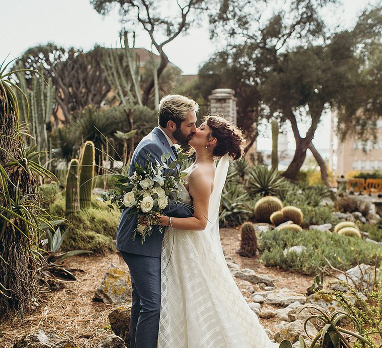 Bride in a strapless Wtoo wedding dress kissing her groom in a blue suit at their Gibraltar Botanic Gardens elopement wedding 