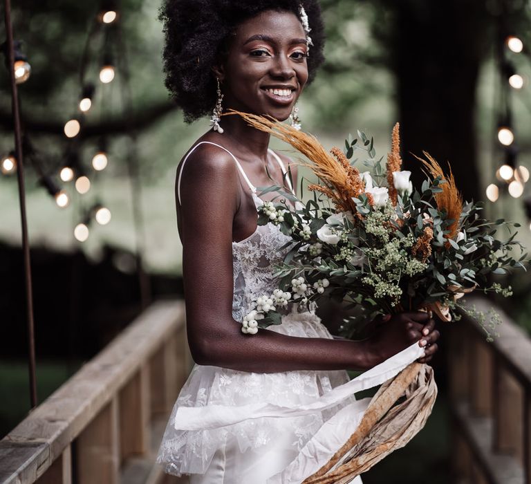 Black bride with afro hair wearing a lace peplum wedding dress holding a orange flower and foliage wedding bouquet tied with ribbon