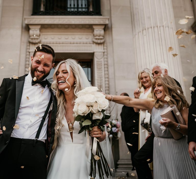 Bride & groom laugh on the steps whilst wedding guests throw confetti 