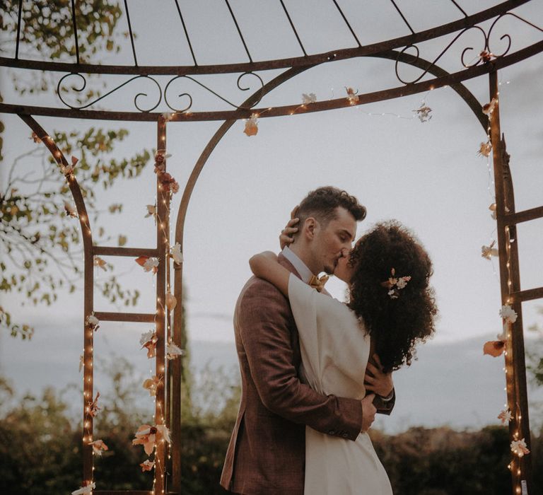 Groom in a brown suit kissing his bride with naturally curly hair under a fairy light decorated pergola 