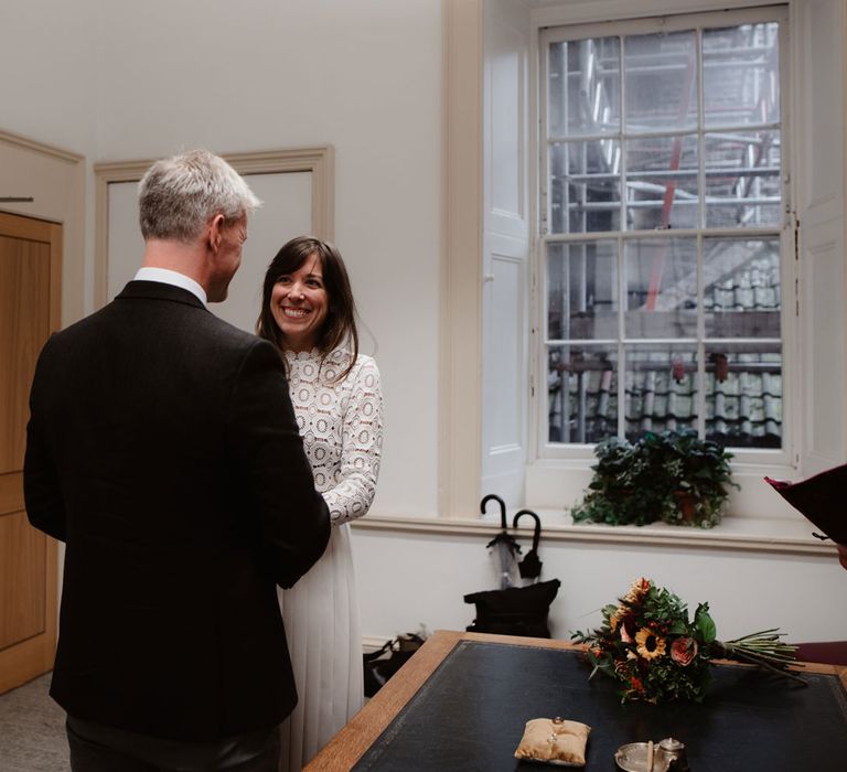 Bride in lace long sleeved Self Portrait dress smiles at groom in brown woollen blazer inside Bristol Registry Office during wedding ceremony