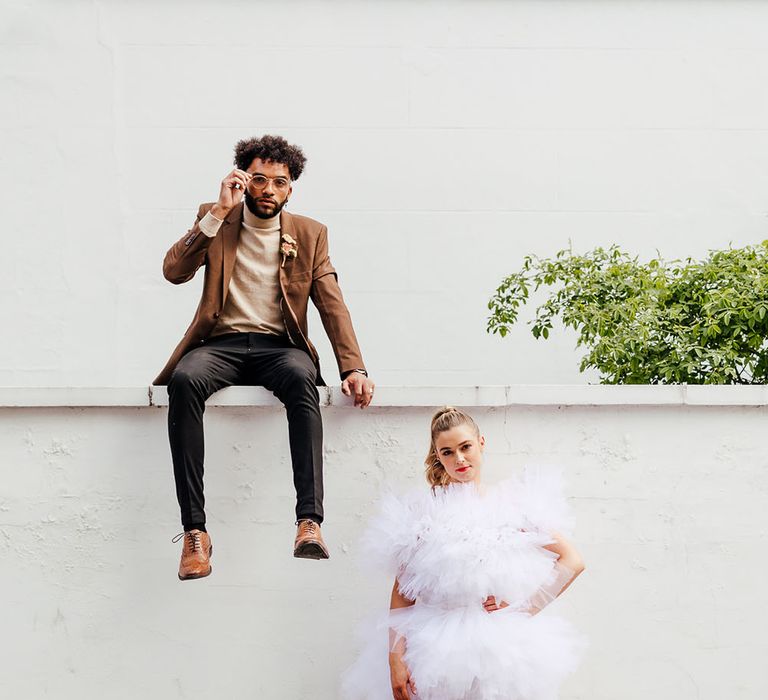 Groom in a beige polo neck and brown blazer sitting on a wall with his bride in a short ruffle wedding dress standing neck to him 