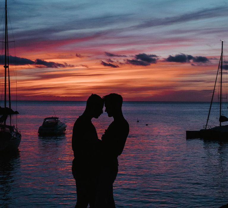 Grooms embrace and kiss whilst the sun sets behind them