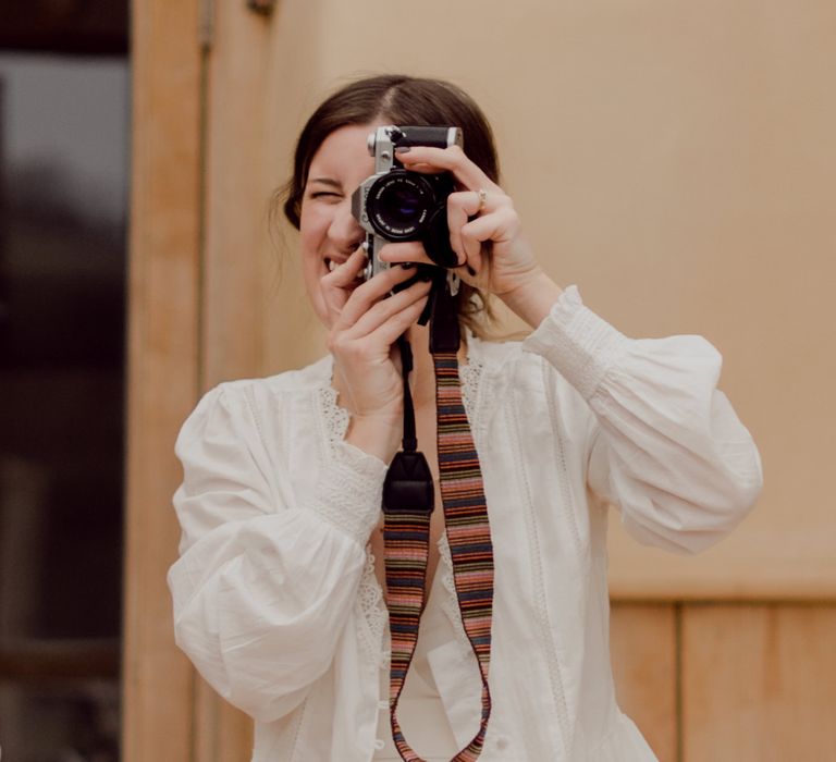 Bride in white blouse and Charlie Brear wedding dress holds film camera up to her eye at garden party wedding in Devon