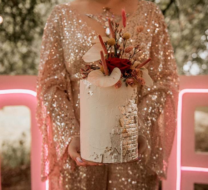 Bride in gold sequinned wedding dress holds white column wedding cake with silver detailing and tropical floral decor at festival themed wedding