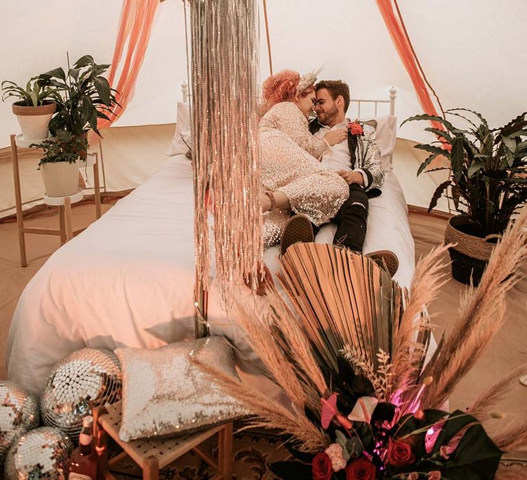 Bride in gold sequin wedding dress and groom in silver sequin suit lie in bed in teepee with festival decor at festival themed wedding