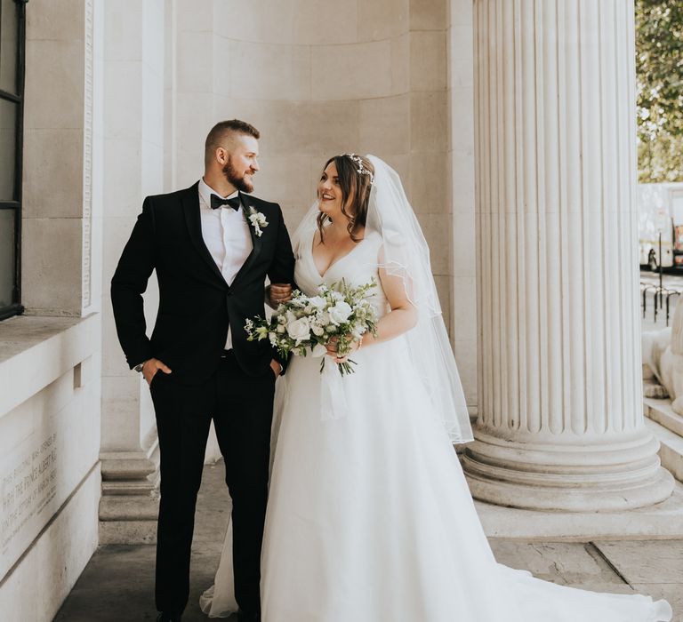 Bride & groom stand beneath white pillars on their wedding day at The Old Marylebone Town Hall