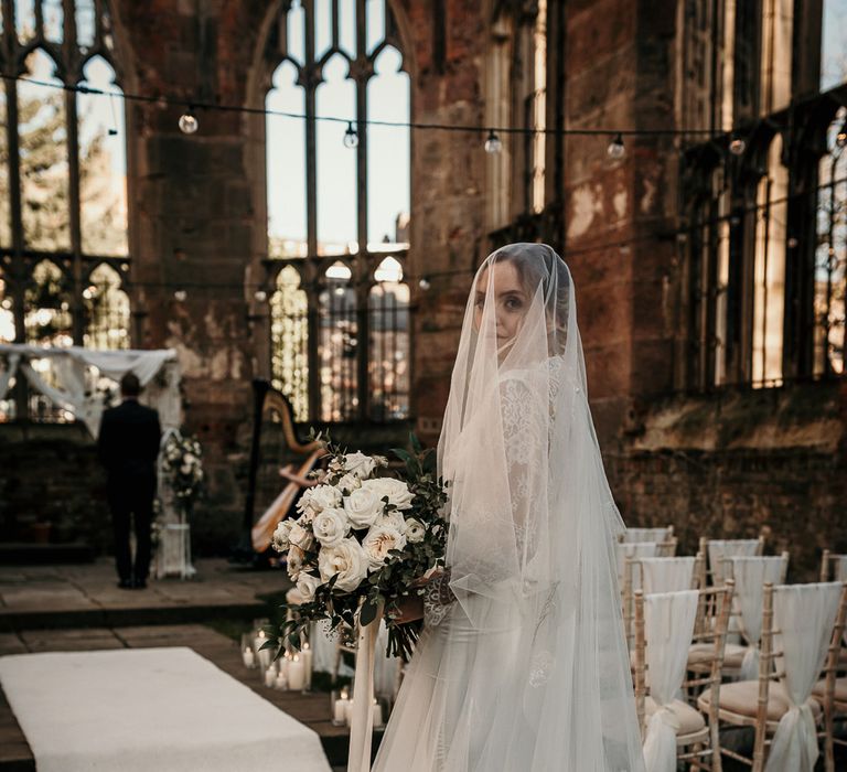 Bride in an appliqué cathedral length veil about to walk down the bombed out church aisle 