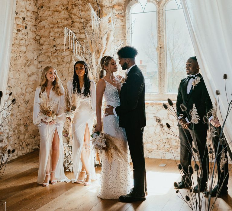 Bride in a appliqué wedding dress with pearl headband standing at the alar with her groom in a tuxedo and bridesmaids in satin dresses 