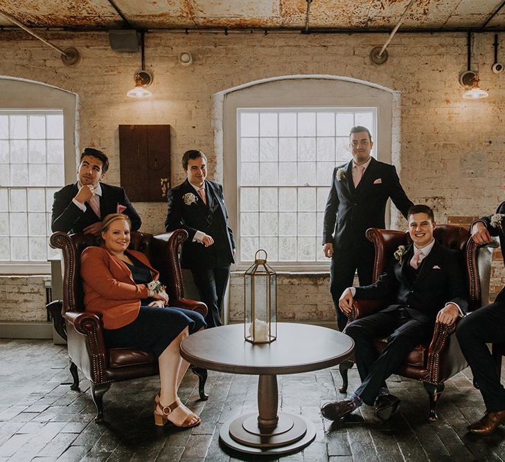 A relaxed group shot of a groom and his party at a wedding. They are gathered around two large leather chairs and a low round table.