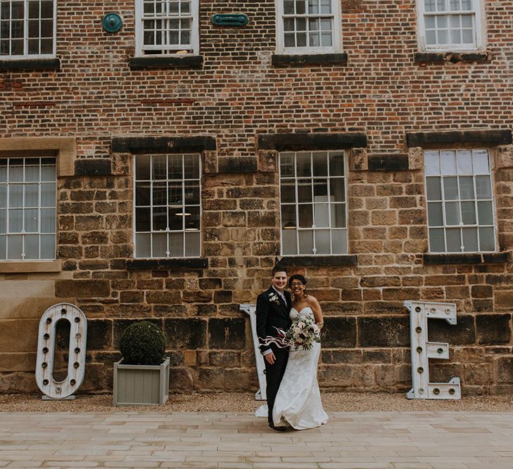 A bride and groom stand outside The West Mill wedding venue and large light up L O V E letters.