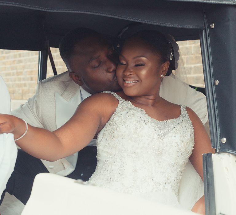 Bride & groom sit within car as groom kisses bride on her cheek and she smiles lovingly