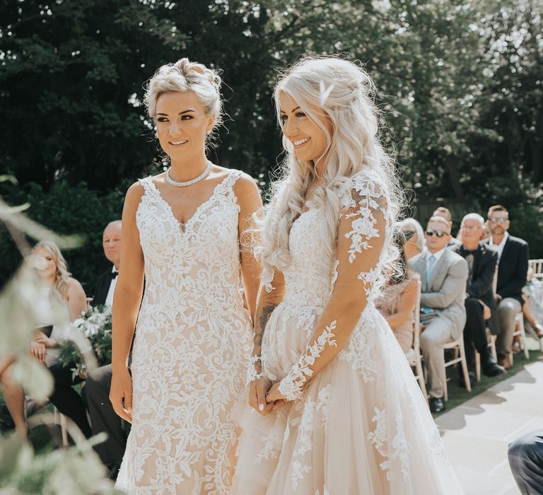 A lesbian couple stand at the alter during their wedding ceremony. They are both blonde. One wears her hair up and the others is long and curly in a half up half down style. 